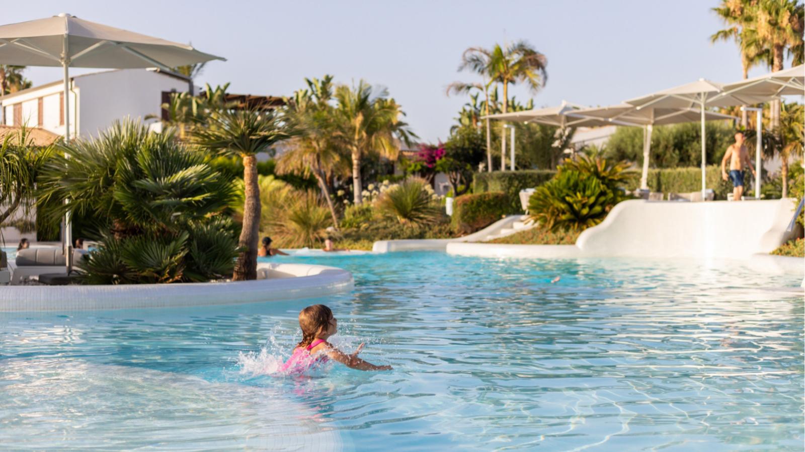 Bambina che gioca in piscina, circondata da palme e sdraio.