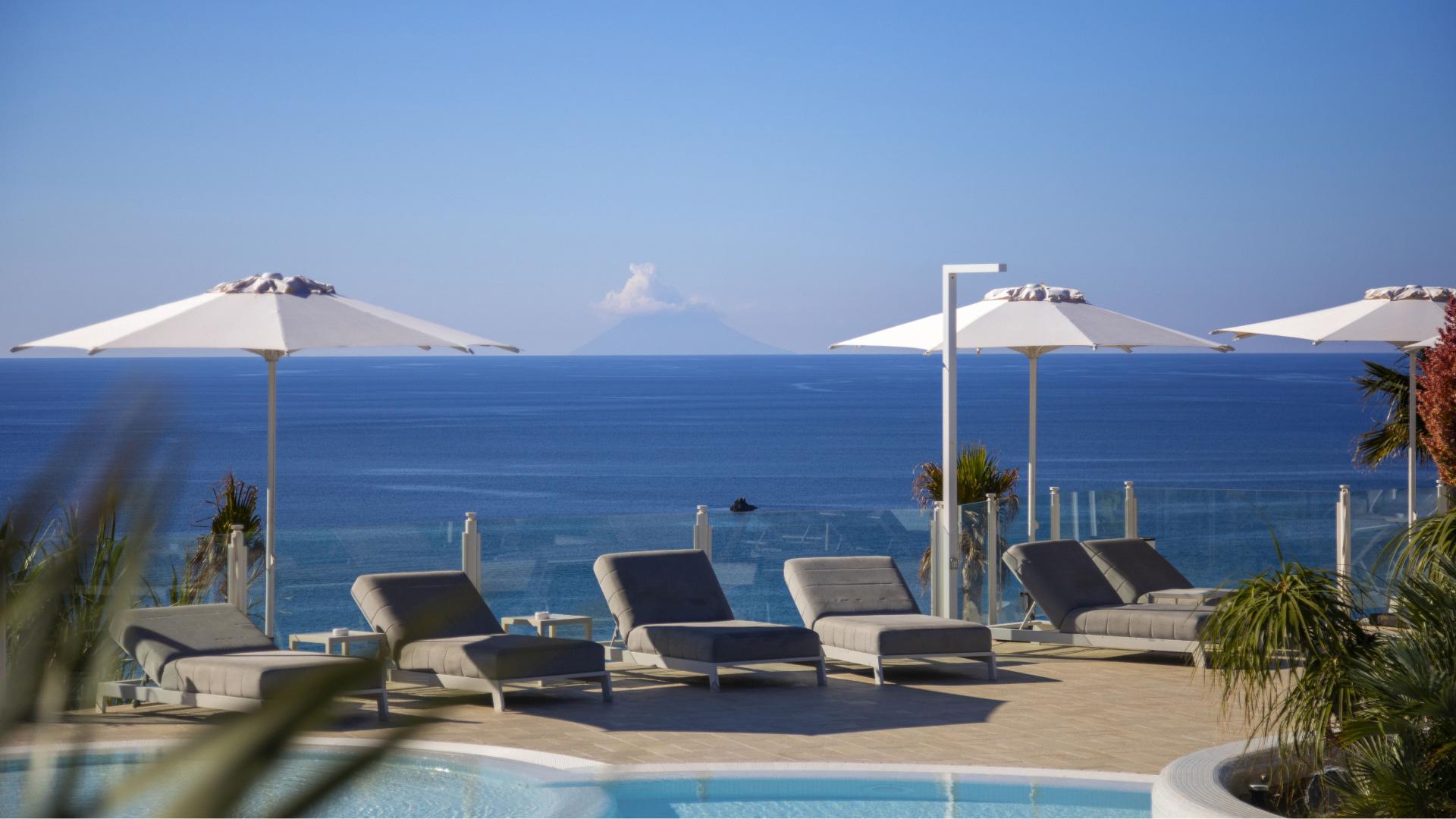 Piscina con lettini e ombrelloni, vista mare e vulcano in lontananza.