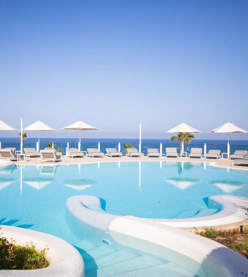 Piscina panoramica con vista mare e lettini sotto ombrelloni bianchi.
