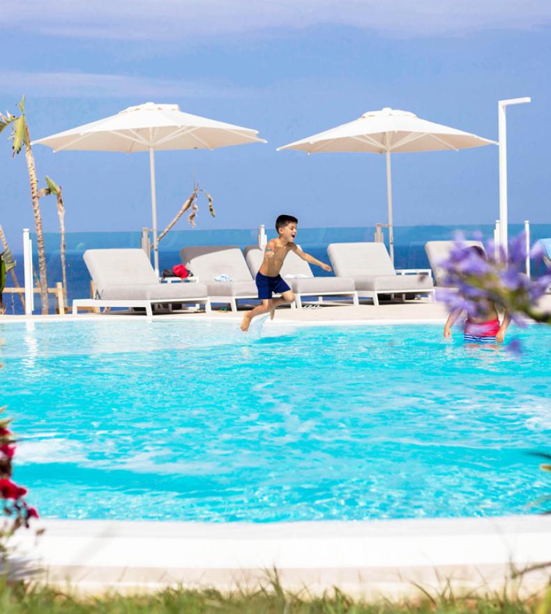 Bambino salta in piscina, ombrelloni e lettini sullo sfondo.